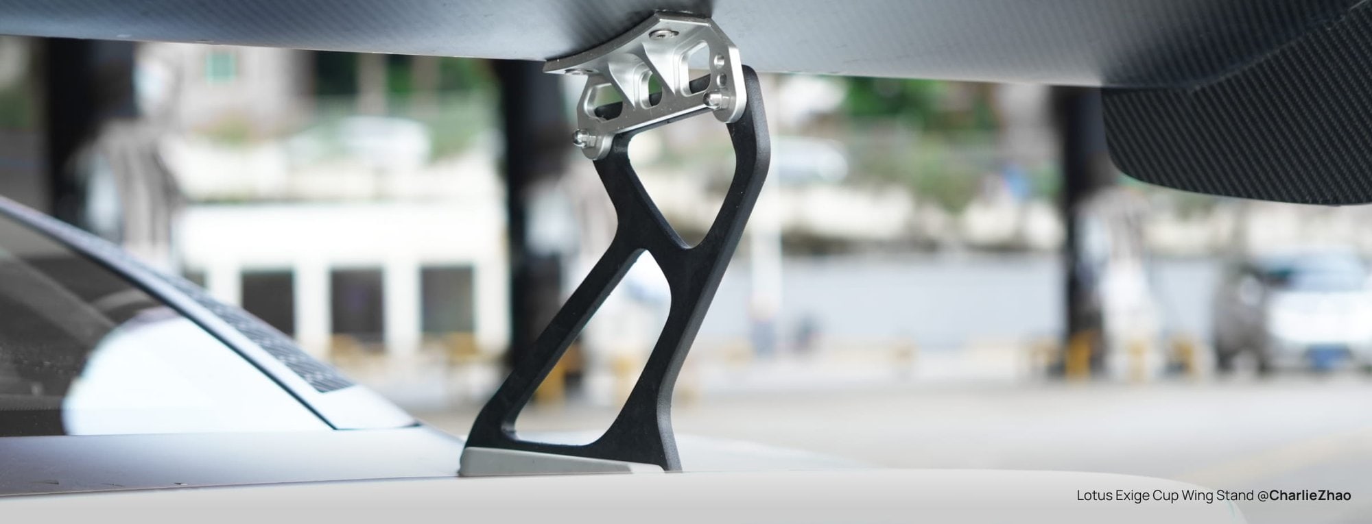 A black PPA-CF wing stand with a silver metal bracket, holding a carbon-fibre rear wing above the white rear deck of a Lotus Exige, outdoors