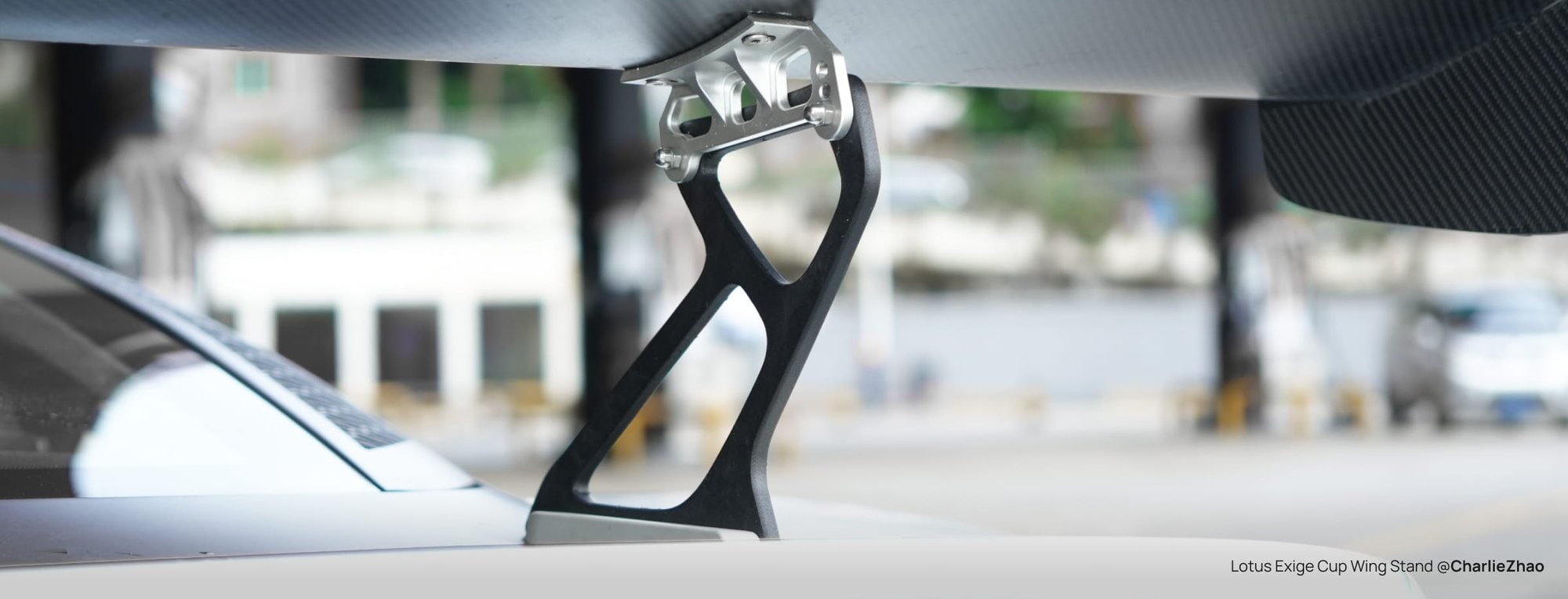 A black PPA-CF wing stand with a silver metal bracket, holding a carbon-fibre rear wing above the white rear deck of a Lotus Exige, outdoors